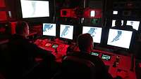 Zwei Marinesoldaten in dunkler Arbeitsuniform sitzen vor einer Konsole mit vier Bildschirmen und weiteren Anzeigen und Knöpfen.