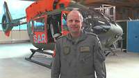 Ein Hubschrauberpilot steht lächelnd vor einem Rettungshubschrauber mit orangefarbenen Aufklebern.