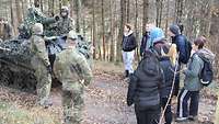 Mehrere Soldaten stehen vor einem gepanzerten Fahrzeug. Eine Gruppe Jugendlicher steht im Halbkreis davor.