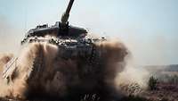 Ein Panzer fährt mit hohem Tempo über trockenes Gelände, Sand und Staub wirbelt auf.
