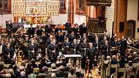 Mehrere Menschen stehen in dunklen Anzügen mit unterschiedlichen Musik-Instrumenten in der Kirche Warnemünde.