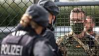 Im Vordergrund ein Polizist und eine Polizistin, die sich durch den Zaun hindurch mit Oberfeldwebel Riedel austauschen.
