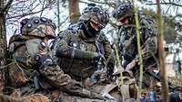 Zwei Soldaten versorgen mit Verbandszeug ihren Kameraden, der im Wald verwundet auf der Erde sitzt.