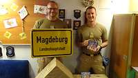 Zwei Soldaten stehen hinter vier großen Weihnachtspaketen. Ein Soldat hält ein Schild mit der Aufschrift Magdeburg in den Händen