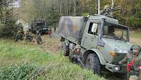 An einem Waldweg stehen zwei Militärfahrzeuge, daneben hocken Soldaten mit Waffen im Anschlag.