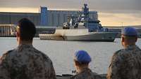 Eine deutsche Korvette läuft in einen Hafen ein, im Vordergrund stehen deutsche UN-Soldaten