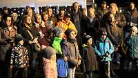 Eine Gruppe Männer und Kinder, alle in Winterkleidung wird vom Adventsfenster angeleuchtet.
