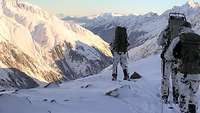 Drei Soldaten mit Skiausrüstung stehen an einem Gebirgsgrat mit dem Blick auf ein gewaltiges Gebirgsmassiv. 