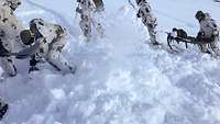 Soldaten graben mit Schaufeln Schnee beiseite, im Hintergrund bereiten Soldaten die Universaltrage für den Transport vor.