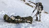 Ein Soldat wird mit der Rettungstrage im Schnee gezogen, während ihm ganz dicht einer folgt.