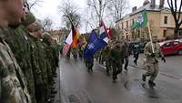 Soldaten mit Flaggen laufen auf einer Straße. Zuschauer auf den Gehsteigen beobachten sie dabei
