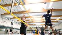 Ein Spieler springt hoch, um den Ball über das Netz zu pritschen, hinter dem Netzt stehen zwei abwehrbereite Spieler.
