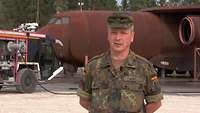 Ein Soldat im Porträt, im Hintergrund steht ein Feuerwehrfahrzeug und ein rostbraunes Übungsflugzeug