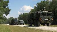 Ein Marschband aus grünen Fahrzeugen steht auf einem Kiesweg am Waldrand. 