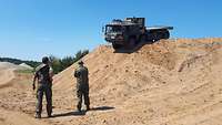 Lastwagen fährt einen Sandhügel hinunter, um Vordergrund stehen zwei Soldaten im Feldanzug.