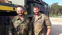 Zwei Soldaten im Feldanzug stehen vor einem Lastwagen in der Sonne.