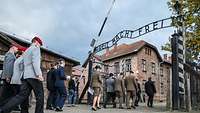 Deutsche und polnische Soldaten laufen durch ein Eingangstor mit dem Schriftzug „Arbeit macht frei“