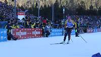 Biathletin auf Ski fährt an einer Zuschauertribüne vorbei.