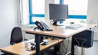 Ein hochgestellter Arbeitstisch mit Monitor, Tastatur und Telefon neben einem Schreibtisch auf Normalhöhe