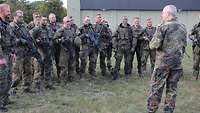 Soldaten mit Waffen und Tarnfarbe stehen im Freien einem General gegenüber und hören ihm zu.