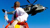 Ein Mann im weißen Pulli dreht sich um, blickt auf einen Helikopter, der über ihm schwebt.