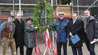 Eine weibliche und fünf männliche Personen, eine davon in Marineuniform, stehen vor einem Richtkranz