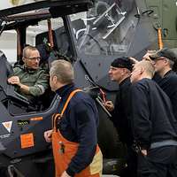 Ein Soldat sitzt im offenen Cockpit eines Hubschraubers und davor stehen sechs zivile Zuhörer.