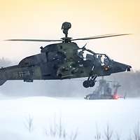 Ein Hubschrauber im Schwebeflug über einer Schneedecke, ein anderer rollt die Startbahn entlang.