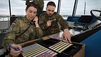 Eine Soldatin und ein Soldat sitzen im Tower eines Flugplatzes nebeneinander und arbeiten.