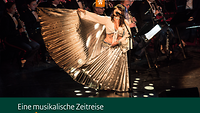 Eine Frau in einem goldglänzenden Kleid steht vor einem Orchester an einem Mikrofon und singt.