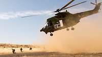 Ein Hubchrauber schwebt über dem Boden und wirbelt Sand auf. Unter ihm hocken Soldaten und sichern die Umgebung