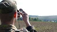 Ein Soldat hält an einem Feldrand einen Kompass in der Hand und peilt einen Punkt in der Ferne an.