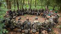 Soldaten sitzen im Gefechtsanzug draußen im Wald im Kreis und tauschen sich aus.