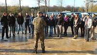 Mehrere Zivilisten stehen im Halbkreis um einen Soldaten, der zu ihnen spricht.