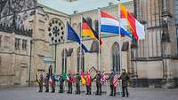 Zehn Soldaten stehen mit kleinen Nationalflaggen vor einer Kirche, hinter ihnen vier große Flaggen.