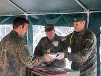 Mittagsverpflegung auf dem Truppenübungsplatz Der Kompiefeldwebel, gekennzeichnet durch eine gelbe Schnur an der rechten Schulter, verteilt Verpflegung an zwei Soldaten.