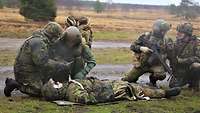 Zwei Soldaten hocken neben einem liegenden verletzten Soldaten, zwei andere sichern die Lage.