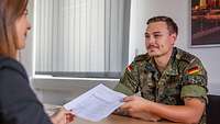 Eine Zivilistin und ein Soldat sitzen sich an einem Tisch gegenüber und sprechen miteinander.