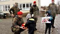 Hockend gibt ein Soldat einem Kleinkind einen roten Kasten, daneben drei Soldaten und ein Junge.
