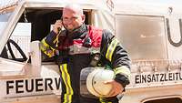 Ein Feuerwehrmann steht vor seinem Feuerwehrauto und hält den Hörer des Funkgerätes in der Hand