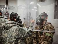 Ein Soldat steht unter einer Plastikhaube, eine amerikanische Soldatin besprüht seine ABC-Maske mit einem Prüfmittel