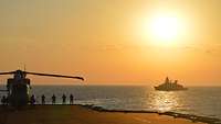 Ein Hubschrauber steht auf dem Flugdeck. Im Hintergrund fährt ein Schiff vor der untergehenden Sonne