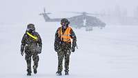 Zwei Bodencrewmitglieder gehen in Schneetreiben die Startbahn entlang, dahinter unscharf ein NH-90.