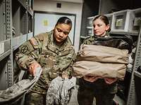 Eine amerikanische Soldatin nimmt Trainingsuniform aus einem Regal, daneben eine deutsche Soldatin mit Kleidung auf dem Arm