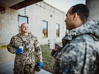 Ein deutscher und ein amerikanischer Soldat halten Trinkbecher in der Hand und unterhalten sich vor einem Gebäude