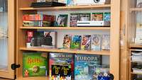 Eine Glasvitrine mit mehreren Spielkonsolen, einem Beamer und Verleih-DVDs