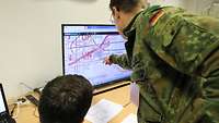 Ein Soldat sitzt und einer steht vor einem PC. Der Stehende zeigt mit dem Finger auf den Bildschirm.