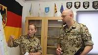 Zwei Soldaten stehen vor einer Tafel, im Hintergrund die deutsche Flagge und Abzeichen 
