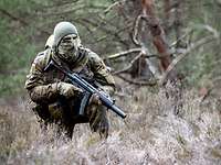 Ein Soldat hockt mit seiner Waffe vor der Brust auf einer Waldlichtung.