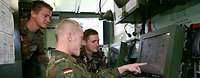 Zwei Soldaten sitzen und einer steht vor einem Bildschirm, auf dem taktische Zeichen zu sehen sind.